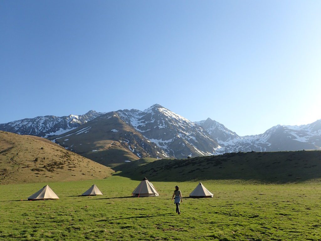 A person walking toward a row of tents located in the middle of a green field under big and snow-covered mountains