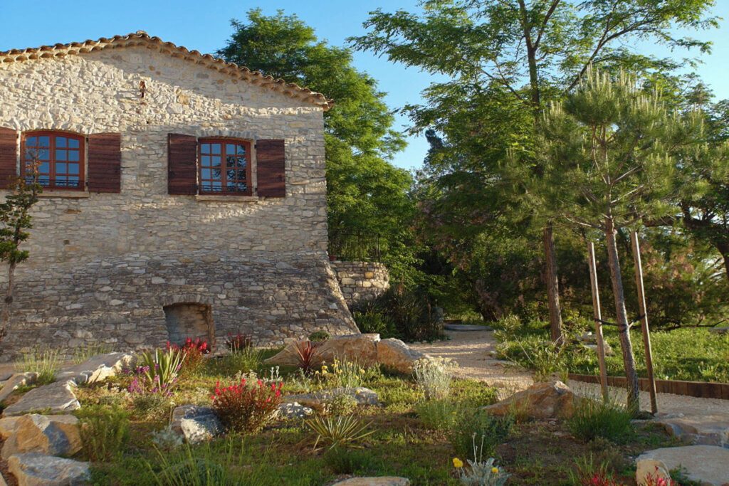A stone house surrounded by a garden path with vibrant wildflowers and green trees under a clear blue sky